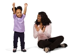 Smiling African American mom sitting on floor clapping while toddler daughter raises her hands smiling. 